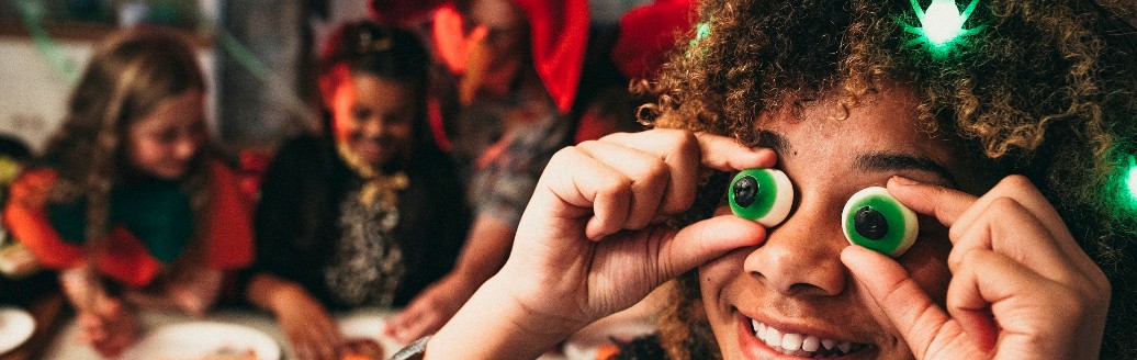 Kinder sind gemeinsam auf einer Halloween-Party und ein Kind macht im Vordergrund des Bildes eine Grimasse mit Augen-Gummibärchen