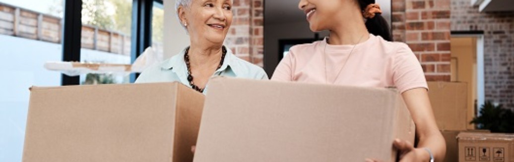 Eine ältere Frau und ein jüngere Teenagerin schauen sich lächelnd an, während sie Umzugskartons packen. 