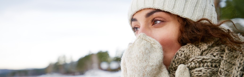 Eine warm eingepackte Frau mit Mütze, Schal und Handschuhen steht draußen in einer verschneiten Landschaft und hält die Hände vor ihr Gesicht, um sich zu wärmen.