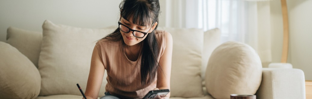 Eine junge Frau sitzt auf einem Sofa, schaut auf Unterlagen auf dem Tisch und macht Notizen, während sie ein Smartphone in der Hand hält.
