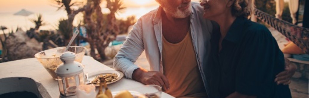Ein älteres Ehepaar (Rentner) genießen ihr Abendessen unter Palmen im Sonnenuntergang. Sie schauen sich läacheldn an.