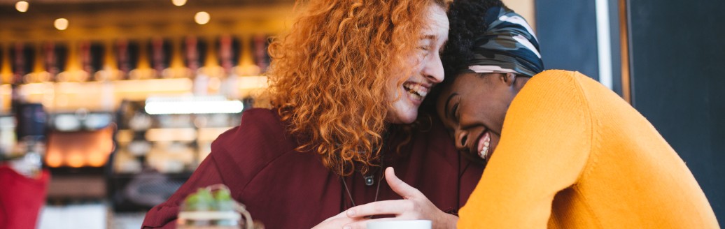 Zwei Frauen sitzen in einem Café und liegen sich lachend in den Armen. 