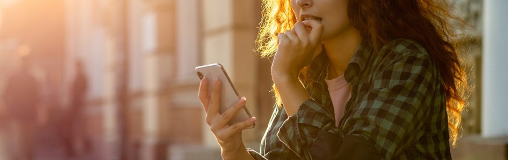 Eine junge Frau sitzt draußen auf einer Steinstufe und schaut gespannt in ihr Smartphone. 