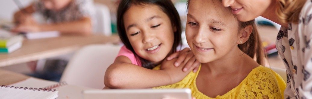 Eine Lehrerin zeigt zwei jungen Schülerinnen etwas auf dem Tablet. Sie sitzen in einem Klassenzimmer.