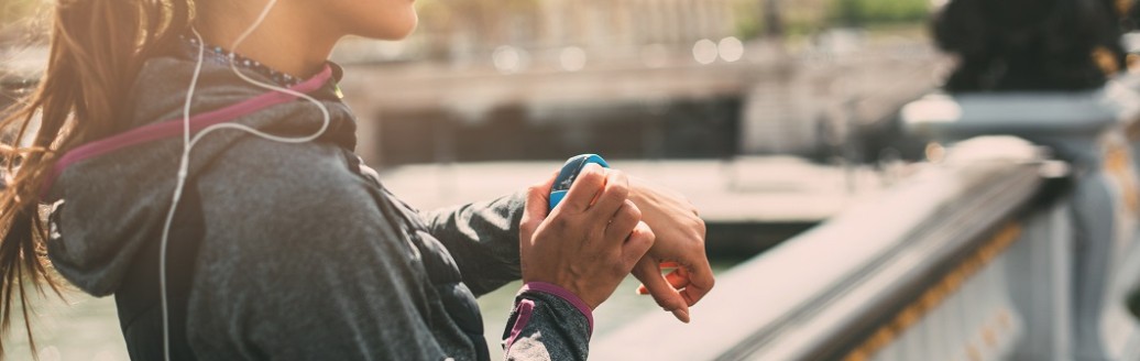 Eine junge Joggerin steht auf einer Brücke und schaut von ihrer Smart Watch hoch. 