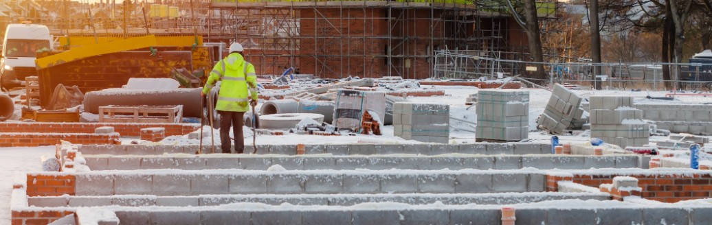Dach einer verschneiten Baustelle. In Ferne kann ein Bauarbeiter in gelber Warn-Schutzkleidung erkannt werden. 