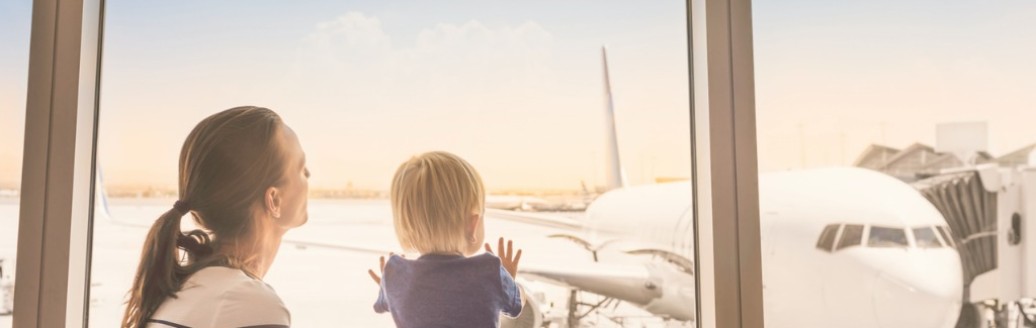 Eine Mutter schaut mit ihrem Kleinkind aus dem Fenster an einem Flughafen. Sie sehen ein Flugzeug, was am Gate steht.