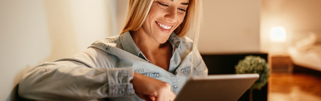 Eine junge Frau lächelt, während sie etwas auf ihr Tablet durchliest. Sie sitzt in ihrem Wohnzimmer auf einer Couch.