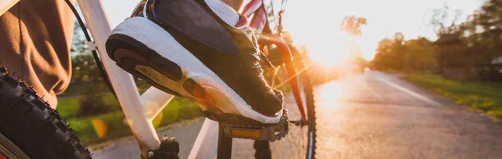 Ein Mann fährt sein Fahrrad im Sonnenuntergang auf einem asphaltierten Weg. Im Fokus stehen seine Sportschuhe.
