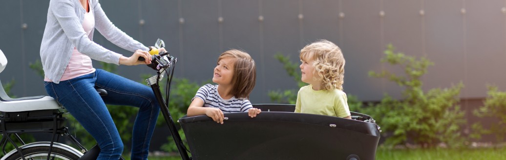 Eine Mutter fährt ein Lastenrad. Vorne im Anhänger sitzen zwei Kinder, die ihre Mutter anschauen. 