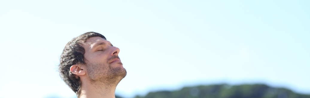 Ein Mann steht draußen mit geschlossenen Augen und hebt das Gesicht leicht in die Sonne, im Hintergrund eine grüne Hügellandschaft.