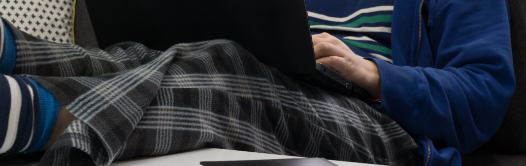 Eine männliche Person (Gesicht nicht zu sehen) liegt auf dem Sofa in Jogginghose und arbeitet am Laptop.