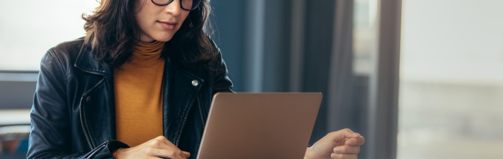Eine Frau sitzt an einem Tisch und schaut auf ihren Laptop. Neben ihr liegen Dokumente. 