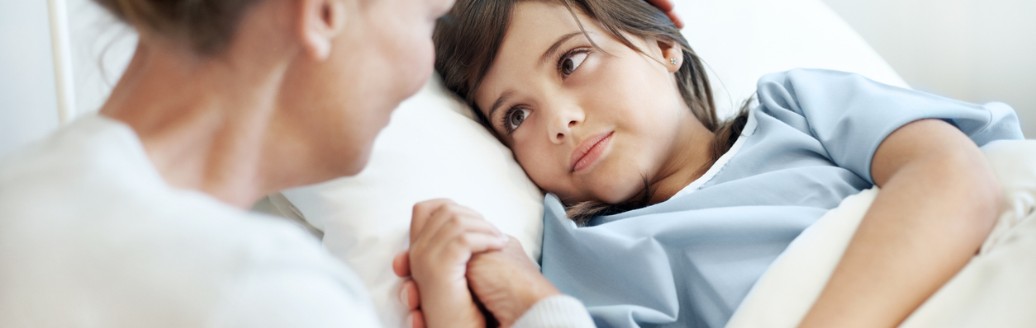 Ein kleiner Junge liegt im Krankenbett. Seine Mutter sitzt daneben und streichelt ihm den Kopf.