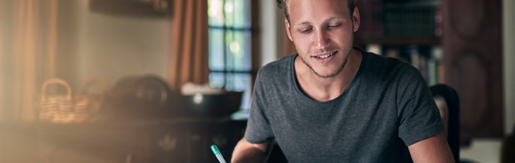 Ein junger Mann arbeitet an etwas mit seinem Laptop. Er sitzt am Schreibtisch. 