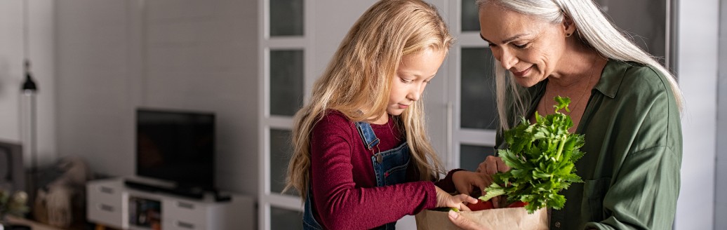 Eine ältere Frau schaut sich mit einem jüngeren Mädchen die Einkäufe in einer Papiertüte an. Sie sitzen zusammen in der Küche.