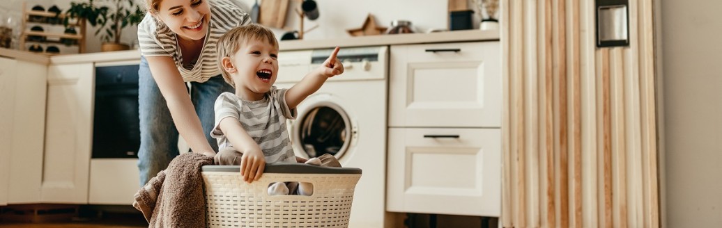 Ein junges Mädchen und ein kleiner Junge albern lachend gemeinsam in der Küche herum. Der Junge sitzt in einem Wäschekorb und wird von dem Mädchen umher geschoben.