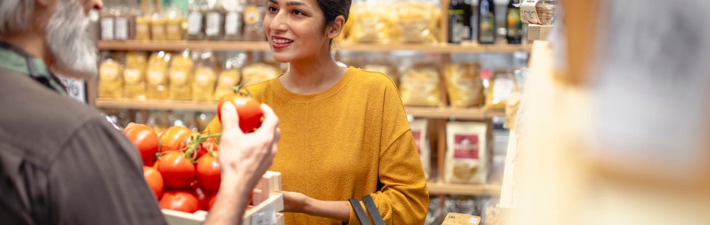 Eine junge Frau lächelt, während sie mit einem älteren Mann in einem Laden spricht und dabei einen Korb mit frischen Lebensmitteln hält. Im Hintergrund sind Regale mit verpackten Produkten zu sehen.