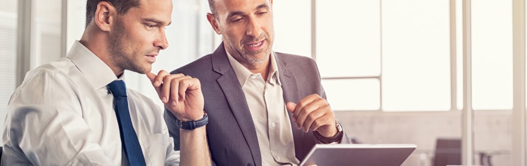 Zwei Business-Männer sitzen zusammen und besprechen etwas am Tablet. 