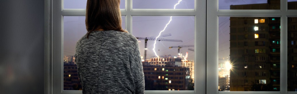 Ein Mädchen sitzt knieend an einem Fenster und schaut die sichtbaren Blitze in der Ferne an. 