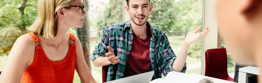 Eine Gruppe von jungen Leuten sitzen zusammen an einem Tisch mit Unterlagen und Laptops. Sie unterhalten sich. 