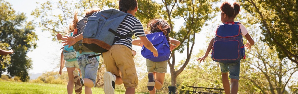 Mehrere Kinder laufen mit ihren Schulrucksäcken über eine Wiese in der Sonne.