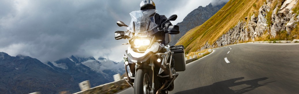 Motorradfahrer fährt auf einer kurvigen Bergstraße, im Hintergrund sind Berge und dunkle Wolken zu sehen.