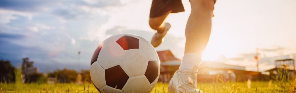 Ein Mann in Fußballklamotten (lediglich der Unterkörper sichtbar) schießt einen Fußball. Drum herum scheint die Sonne. 