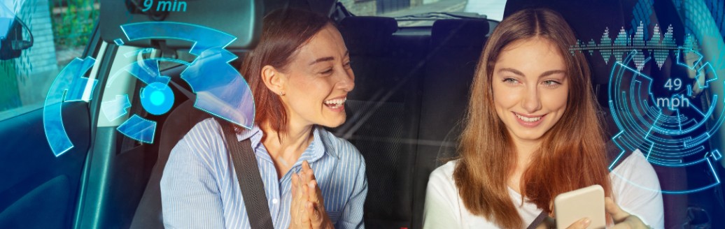 Zwei Freundinnen sitzen zusammen vorne im Auto und schauen in ein Smartphone.