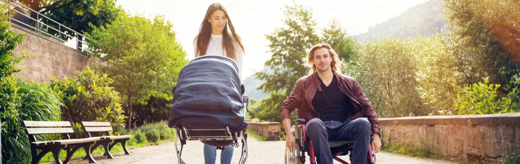 Eine Frau schiebt einen Kinderwagen, während ein Mann daneben in einem Rollstuhl sitzt. Sie spazieren gemeinsam einen Weg entlang. 