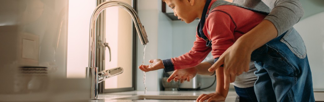 Kleiner Junge steigt auf einem Stuhl, um sich die Hände zu waschen während seine Mutter ihm hilft.