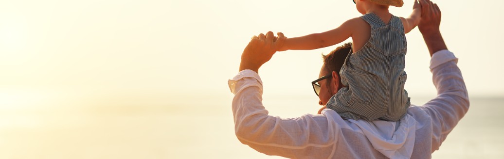Ein Mann steht am Strand am Meer und trägt sein Kleinkind auf den Schultern. 