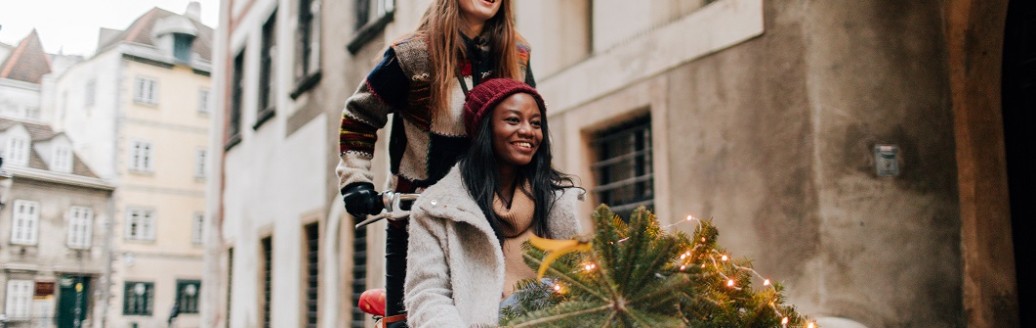 Zwei Freundinnen sind zusammen mit einem Lastenrad durch die Straßen unterwegs. Eine Person fährt das Rad, die andere Person sitzt drin. Sie transportieren einen kleinen Nadelbaum.