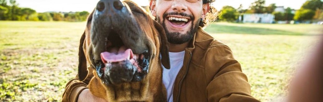 Ein junger Mann und ein Hund schauen gemeinsam in eine Kamera (Selfie-Ansicht). Sie sind draußen im Grünen.