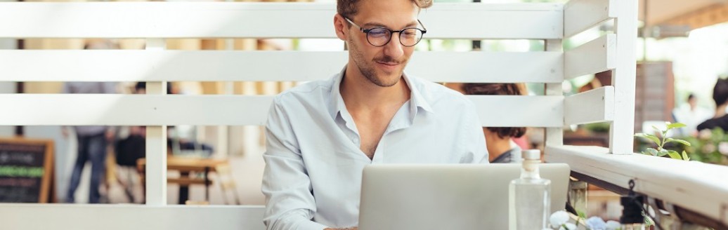 Ein junger Mann sitzt an seinem Laptop und einem Kaffee in einem Café. 