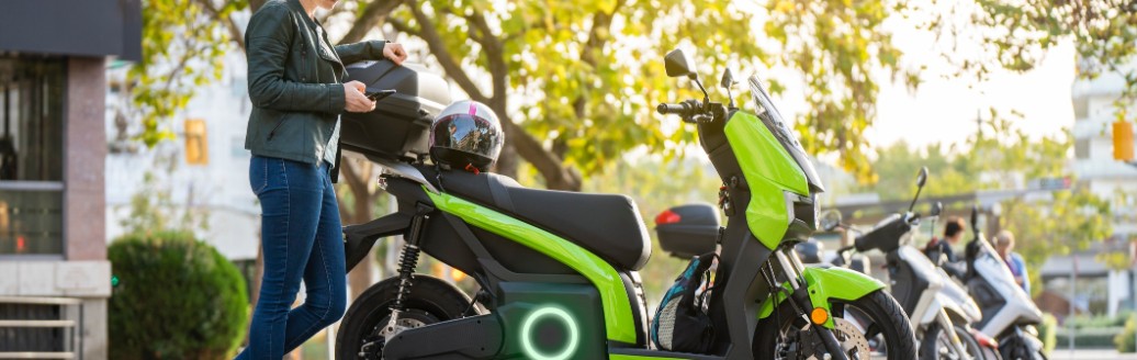 Eine blonde Frau steht neben ihrem elektrischen Motorrad und hält ihren Helm in der Hand. 