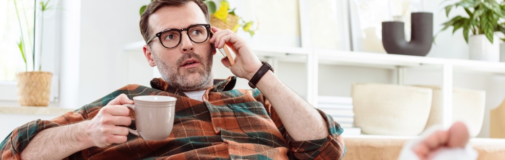 Ein Mann mit Brille sitzt auf einem Sofa, telefoniert und hält eine Tasse. Sein Bein ist in Gips und hochgelegt.
