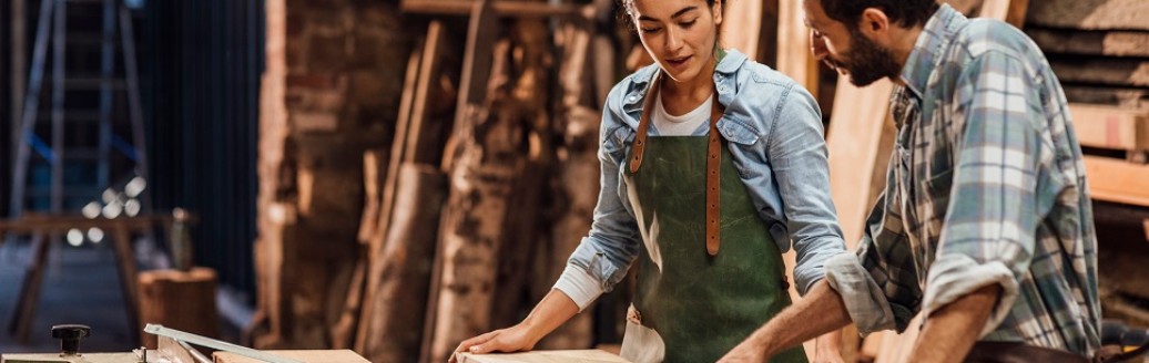 Eine junge Frau und ein junger Mann schneiden zusammen in einer Werkstatt Holz zu.