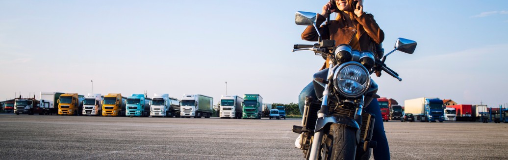 Frau sitzt mit Helm auf einem Motorrad auf einem großen Parkplatz, im Hintergrund stehen Lkw.