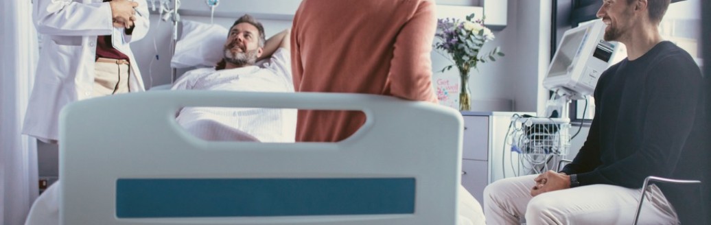 Ein männlicher Patient liegt in einem Krankenhausbett. Rechts von ihm steht eine weibliche Ärztin, eine Frau sitzt am Fußende des Bettes und ein Mann daneben auf einem Stuhl. 