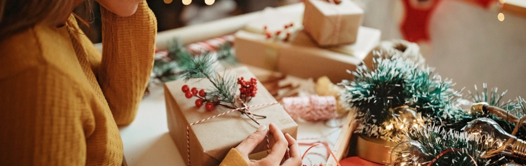 Ein Mädchen sitzt nachdenklich am Schreibtisch und begutachtet ein eingepacktes Weihnachtsgeschenk. Um sie herum ist Weihnachtsdekoration und andere eingepackte Weihnachtspäckchen. 