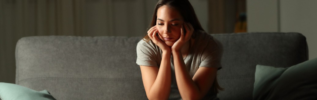 Eine junge Frau sitzt in ihrem Wohnzimmer ohne Strom und schaut ernüchternd auf drei leuchtende Kerzen auf dem Wohnzimmertisch. 