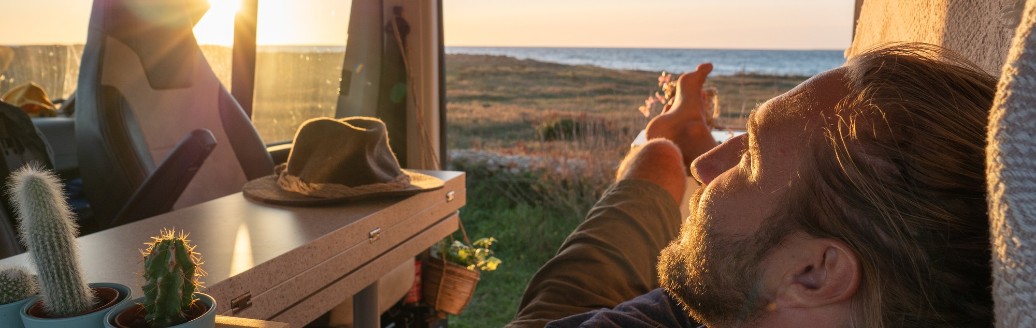 Ein junger Mann liegt in seinem ausgebauten Campervan mit Sonne im Hintergrund.