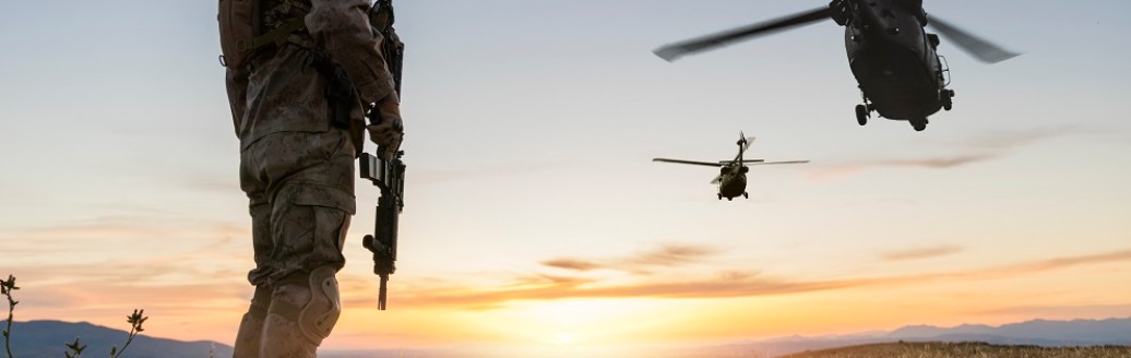 Ein Soldat steht bewaffnet auf einem Hügel schaut in Richtung wegfliegender Militärhubschrauber. 