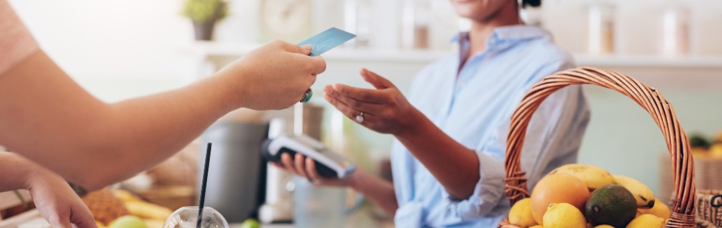 Eine Hand hält einer Frau eine Bankkarte hin. Auf dem Tresen steht ein Smoothie und die Frau hinter dem Tresen kassiert ab. 