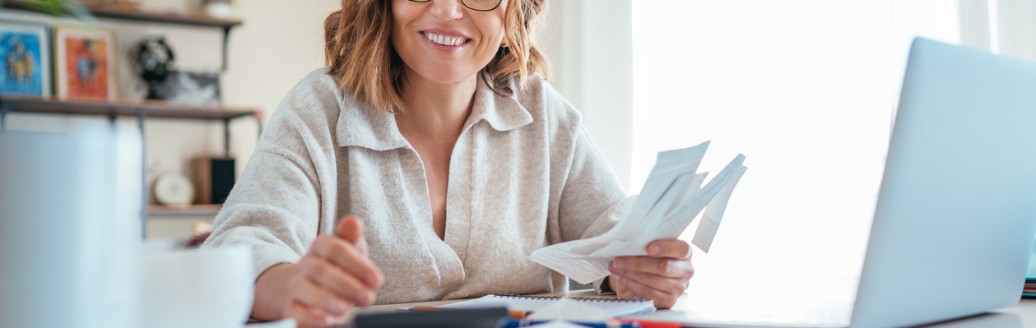 Frau mit Brille sitzt lächelnd an einem Schreibtisch, hält Belege in der Hand und arbeitet mit Taschenrechner und Laptop.