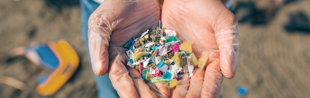 Zwei Hände halten kleine Teile aus Mikroplastik. Im Hintergrund ist ein Strand zu erkennen. 