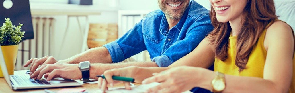 Ein Mann und eine Frau sitzen nebeneinander und unterhalten sich lachend, während sie an Unterlagen und am Laptop arbeiten. 