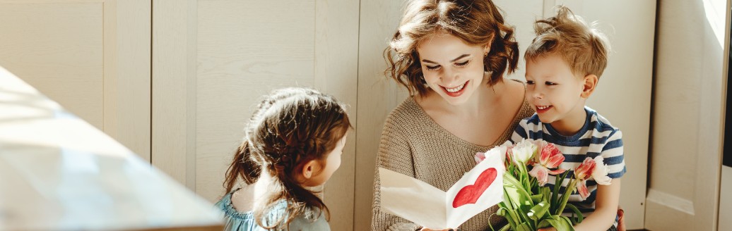 Eine Mutter sitzt auf dem Boden in der Küche und liest ihrem beiden Kindern ein Buch vor. 