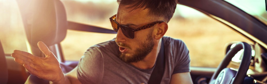 Ein Mann mit Sonnenbrille schaut verwundert und Schultern zuckend um sich im Auto.  